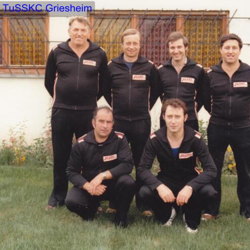 Sechsköpfige Gruppe von Männern in schwarzen Trainingsanzügen, posierend vor einem Gebäude mit bunten Fenstern.