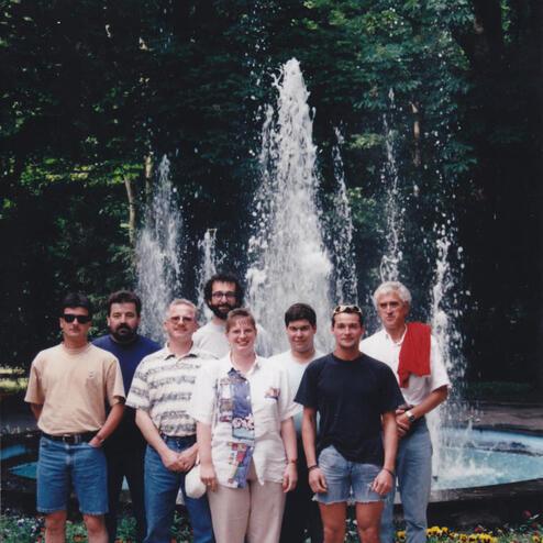 Gruppe von sieben Personen posiert vor einem Wasserfall in einem parkähnlichen Umfeld, umgeben von grüner Vegetation.