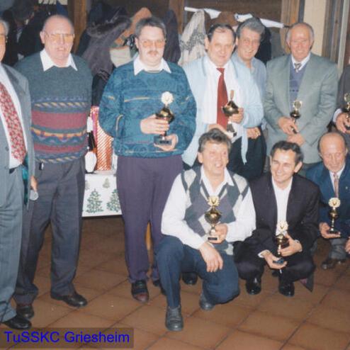 Gruppe von Männern mit Pokalen in einem festlichen Raum, einige stehen, andere sitzen. Weihnachtliche Dekoration im Hintergrund.