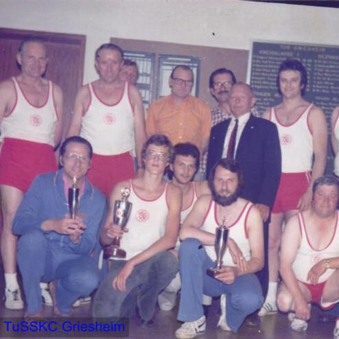 Eine Gruppe von zehn Männern in Sportkleidung posiert lächelnd mit Pokalen in einem Raum.