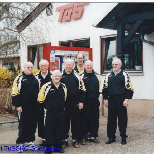 Gruppe von sieben Männern in Sportanzügen vor einem Vereinsgebäude mit "TuS"-Schild und Informationsbrett.