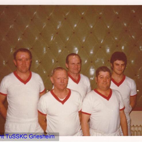 Fünf Männer in weißen T-Shirts mit roten Akzenten stehen vor einer grünen Wand mit samtiger Oberfläche.