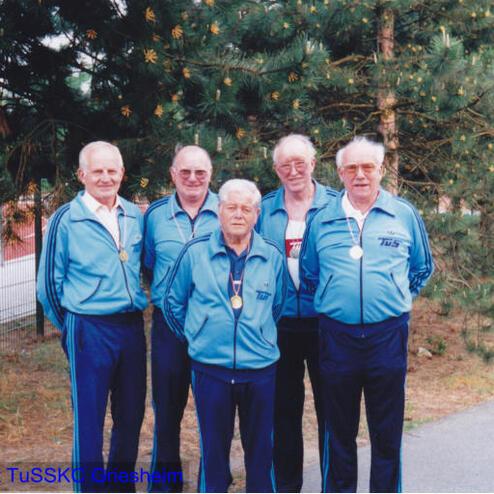 Fünf ältere Männer in blauen Sportanzügen mit Medaillen stehen vor einer Kiefer und lächeln in die Kamera.
