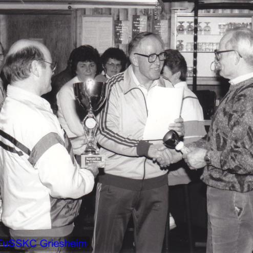 Zwei Personen schütteln sich die Hände, während eine dritte Person einen Pokal hält, in einem festlichen Vereinsraum.