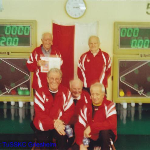 Sechs Männer in roten Trainingsjacken posieren vor einer Sportanzeigetafel und einer Uhr in einem Wettkampfsaal.