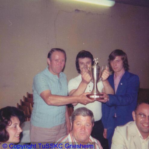 Vier Männer präsentieren gemeinsam einen Pokal in einem festlichen Raum, während zwei weitere Personen am Tisch sitzen.