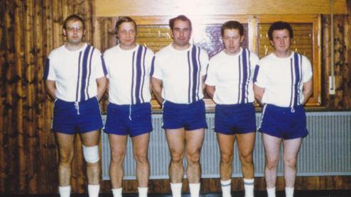 F&uuml;nf sportlich gekleidete M&auml;nner in wei&szlig;en und blauen Trikots posieren gemeinsam vor einer Holzvert&auml;felung.