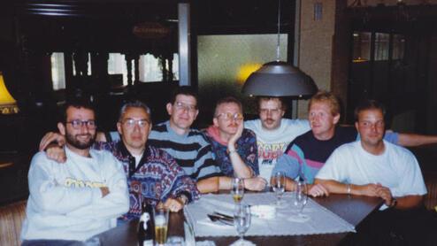 Gruppe von sechs Männern an einem Tisch in einem Restaurant, umgeben von Gläsern und einer Lampe im Hintergrund.