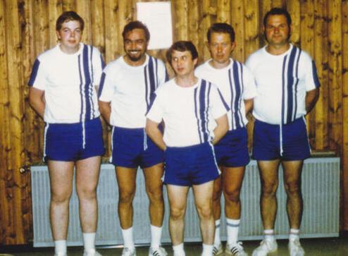 F&uuml;nf M&auml;nner in Sportbekleidung mit blauen Shorts und wei&szlig;en Shirts posieren vor einer Holzvert&auml;felung.