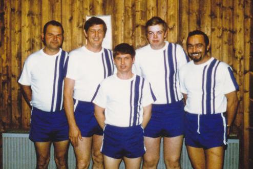 Fünf Männer in Sportbekleidung mit weißen T-Shirts und blauen Shorts stehen vor einer Holzwand.