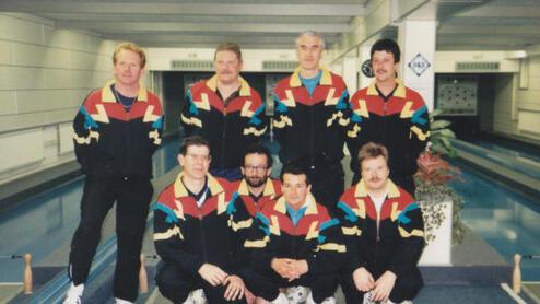Foto einer Gruppe von acht männlichen Sportlern in bunten Trainingsanzügen, auf einer Kegelbahn posierend.