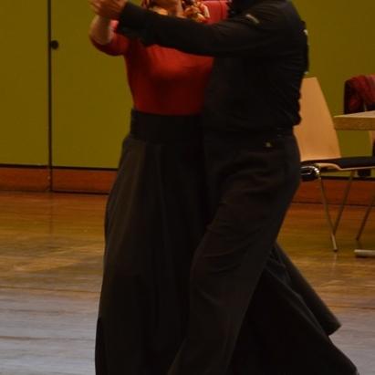 Tanzendes Paar im Vordergrund, Frau in rotem Oberteil und schwarzem Rock, Mann in schwarzer Kleidung, Tanzfl&auml;che im Hintergrund.