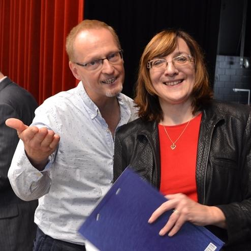 Zwei Personen stehen nah beieinander und l&auml;cheln, w&auml;hrend eine dritte Person im Hintergrund besch&auml;ftigt ist.