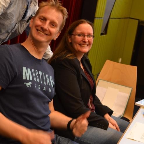 Zwei l&auml;chelnde Personen sitzen an einem Tisch, ausgestattet mit Technik und Notizen in einem Veranstaltungsraum.