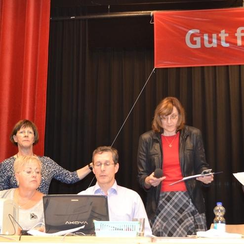 Sechs Personen an einem Tisch, eine h&auml;lt ein Mikrofon, w&auml;hrend eine andere auf einen Computerbildschirm schaut.