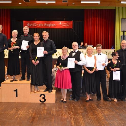 Teilnehmer eines Tanzwettbewerbs stehen auf einem Siegerpodium mit Urkunden und Blumen in der Hand.