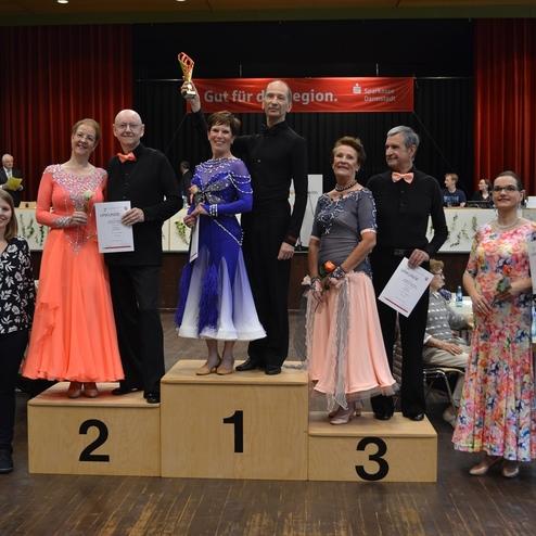 Sieben tanzende Paare stehen auf einem Podium bei einer Tanzveranstaltung, mit Urkunden und Troph&auml;en in den H&auml;nden.