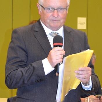 Mann in Anzug h&auml;lt Mikrofon und Dokumente, blickt in die Kamera, Hintergrund mit gr&uuml;nem Wand und Tisch.