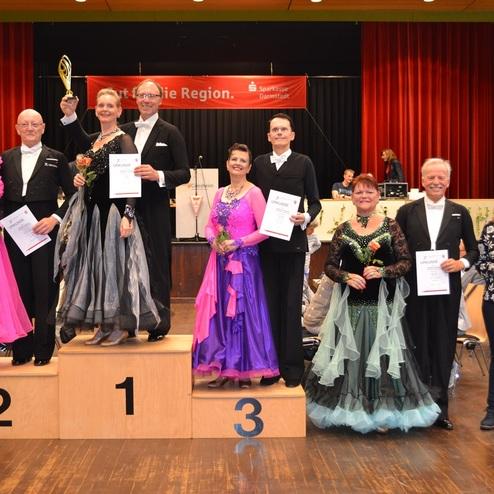 Paarweise stehende T&auml;nzer in eleganter Abendkleidung auf dem Siegertreppchen, Urkunden in der Hand, gemeinsam jubelnd.