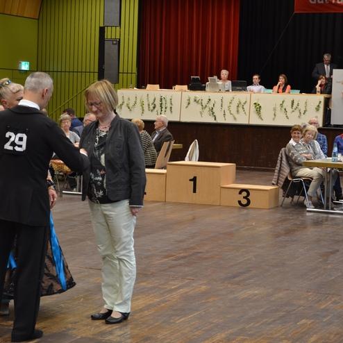 Tanzpaar mit Nummer 29 &uuml;bergibt Pokal an eine Frau in einem festlichen Raum, Zuschauer sitzen an Tischen im Hintergrund.