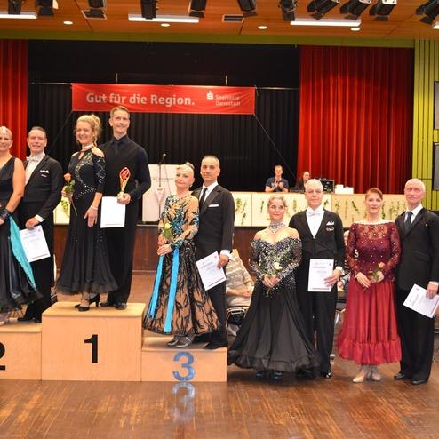 Siegerpaar und Finalisten eines Tanzwettbewerbs stehen auf dem Podest in festlicher Kleidung, mit Urkunden und Blumen.