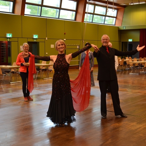 Tanzende Paare in festlicher Kleidung auf einer Holzbodenfl&auml;che, umgeben von St&uuml;hlen und Tischen in einem Saal.