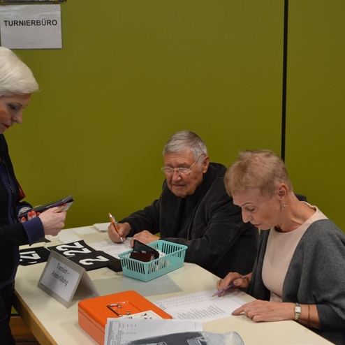 Drei Personen am Tisch im Turnierb&uuml;ro, eine Person steht, zwei sitzen und pr&uuml;fen Anmeldungen auf einem gr&uuml;nen Hintergrund.