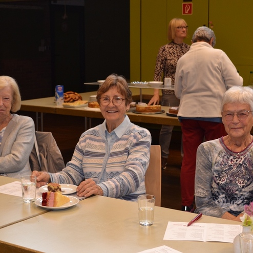 Drei &auml;ltere Frauen sitzen an einem Tisch mit belegten Tellern und Wassergl&auml;sern, im Hintergrund verschiedene Kuchen.