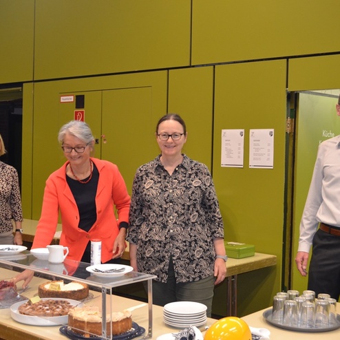 Vier Personen stehen an einem Tisch mit verschiedenen Kuchen und Getr&auml;nken in einem hellgr&uuml;nen Raum.