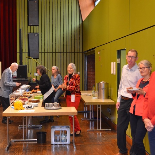 Gruppierung von Personen bei einem Buffet in einem Saal, mit Kuchen und Getr&auml;nken auf Tischen. Farbig gestaltete W&auml;nde.