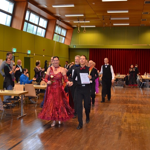 Tanzpaar in festlicher Kleidung mit einer Urkunde, umgeben von applaudierenden Zuschauern in einem gro&szlig;en Saal.