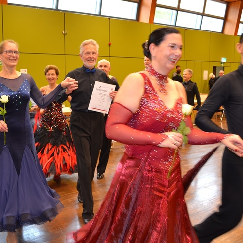 Tanzpaare in festlichen Kleidern, l&auml;chelnd und mit Urkunden in den H&auml;nden, gehen durch einen Saal mit gr&uuml;nem Hintergrund.