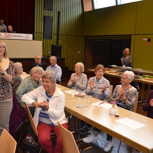 Gruppe &auml;lterer Menschen sitzt an Tischen in einem Veranstaltungsraum und applaudiert, B&uuml;hnenbereich im Hintergrund sichtbar.