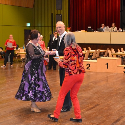 Drei Menschen in festlicher Kleidung unterhalten sich in einem Saal mit St&uuml;hlen und Tischen im Hintergrund.