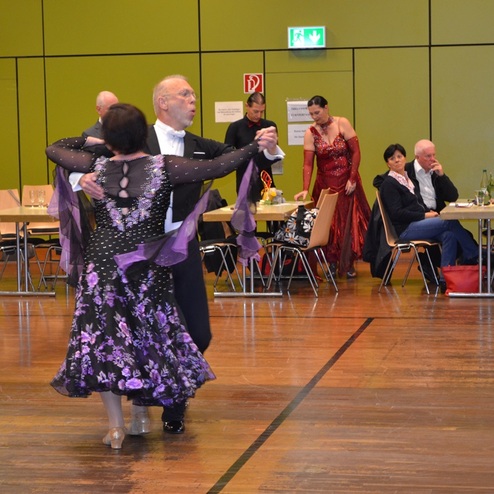 Tanzpaar in eleganter Kleidung auf einer Tanzfl&auml;che, Zuschauer in Abendgarderobe im Hintergrund.
