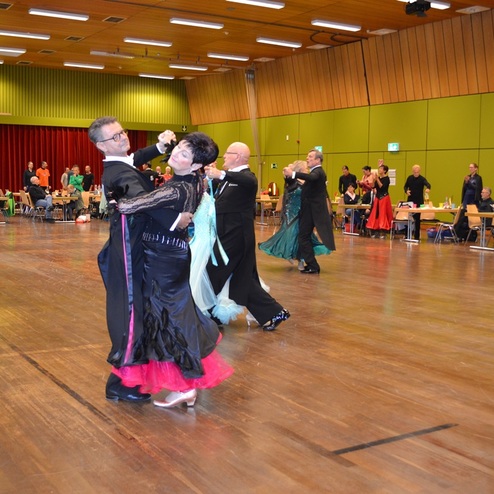 Tanzpaare in festlicher Kleidung tanzen auf einer Turnierfl&auml;che, Zuschauer sitzen an Tischen im Hintergrund.