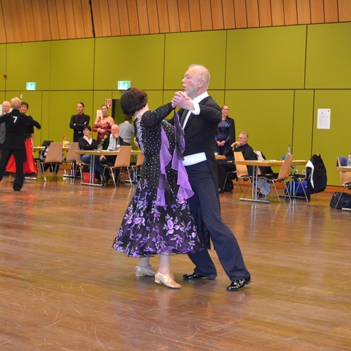 Tanzpaar in eleganter Kleidung f&uuml;hrt einen Tanz auf einer Fl&auml;che, umgeben von Zuschauern und weiteren T&auml;nzern.