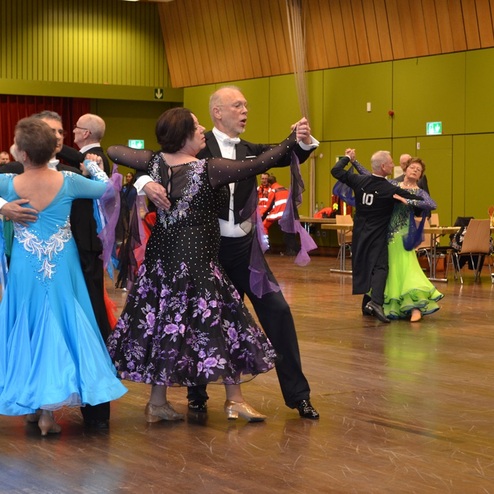 Tanzpaare in eleganten Kleidern f&uuml;hren einen Gesellschaftstanz auf einer Holzfl&auml;che mit Zuschauern im Hintergrund auf.
