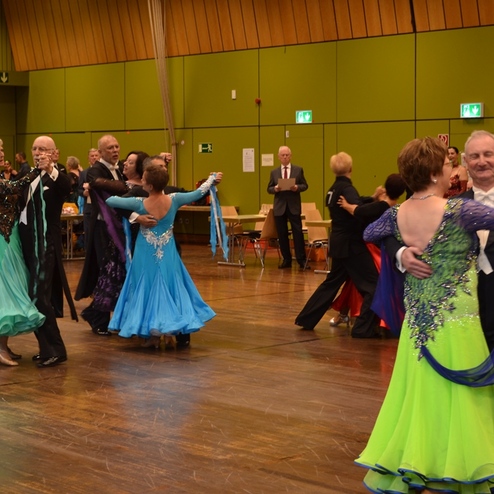 Tanzende Paare in festlicher Kleidung auf einer Tanzveranstaltung in einem hellen Saal mit Holzfu&szlig;boden.