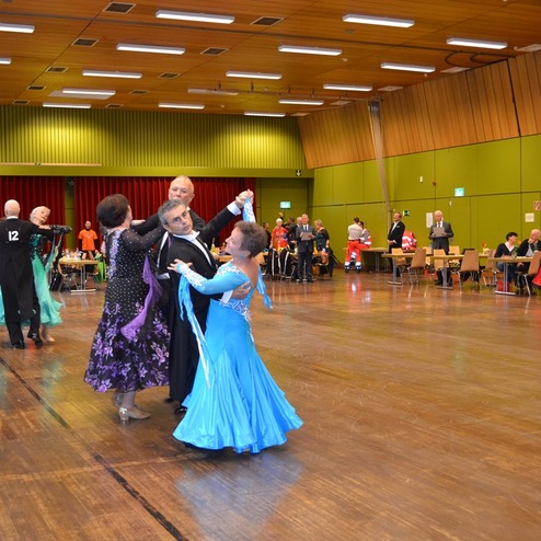 Tanzpaare in festlicher Kleidung tanzen in einer gro&szlig;en Halle, Zuschauer sitzen an Tischen im Hintergrund.