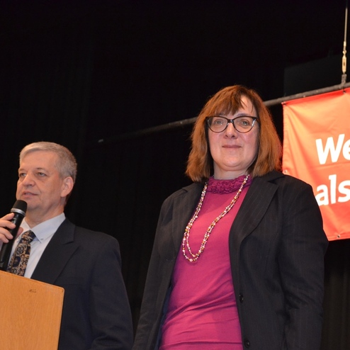 Zwei Personen stehen auf einer B&uuml;hne, ein Mann spricht ins Mikrofon, eine Frau l&auml;chelt daneben. Im Hintergrund ein Plakat.