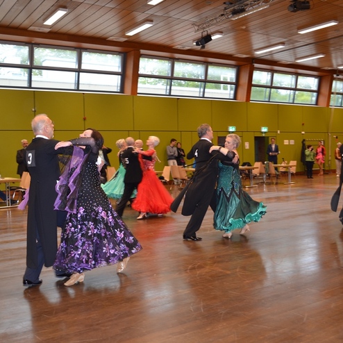 Tanzpaare in eleganten Outfits f&uuml;hren in einem Saal eine Tanzauff&uuml;hrung auf einem Holzfu&szlig;boden auf.