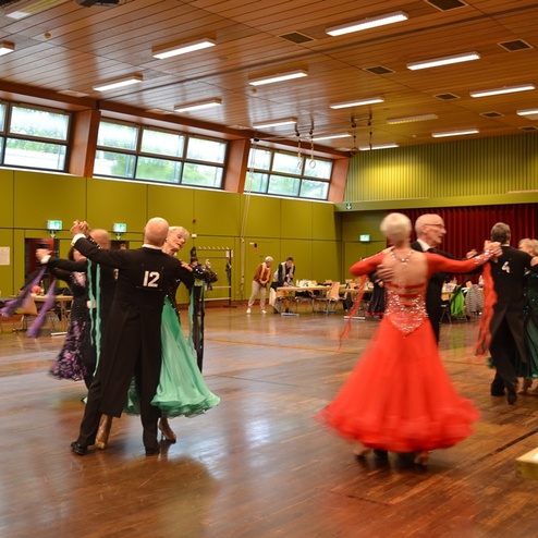 Tanzende Paare in eleganter Abendgarderobe bei einer Tanzveranstaltung in einer gro&szlig;en Halle mit gr&uuml;nen W&auml;nden.