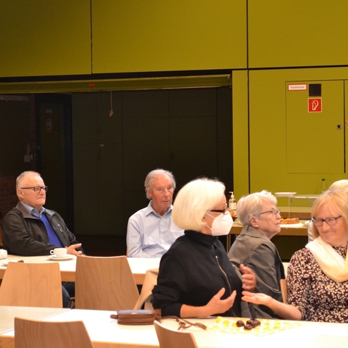 Eine Gruppe &auml;lterer Menschen sitzt an Tischen in einem gro&szlig;en Raum mit gelbgr&uuml;nen W&auml;nden.