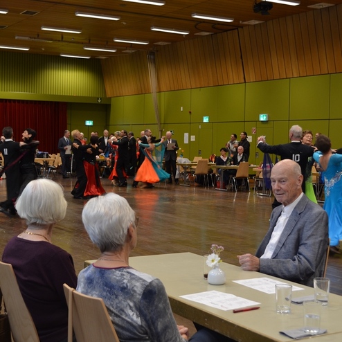 Tanzende Paare in Festkleidung auf einer Veranstaltung, im Vordergrund sitzen Zuschauer an Tischen.