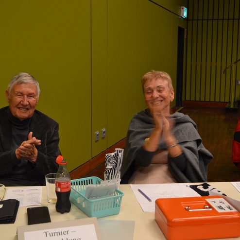 Zwei &auml;ltere Personen sitzen an einem Tisch, l&auml;cheln und klatschen. Auf dem Tisch liegen Unterlagen und Getr&auml;nke.