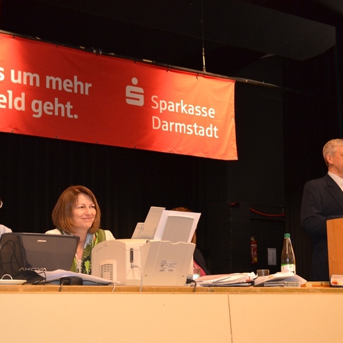 Drei Personen sitzen an einem Tisch, w&auml;hrend ein Mann am Rednerpult vor einem Banner der Sparkasse Darmstadt spricht.