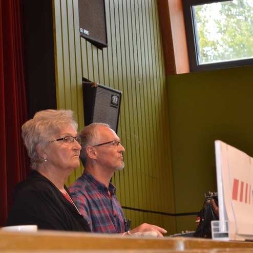 Zwei Personen sitzen an einem Tisch in einem Veranstaltungsraum mit Mikrofonen und Rednerpulten.