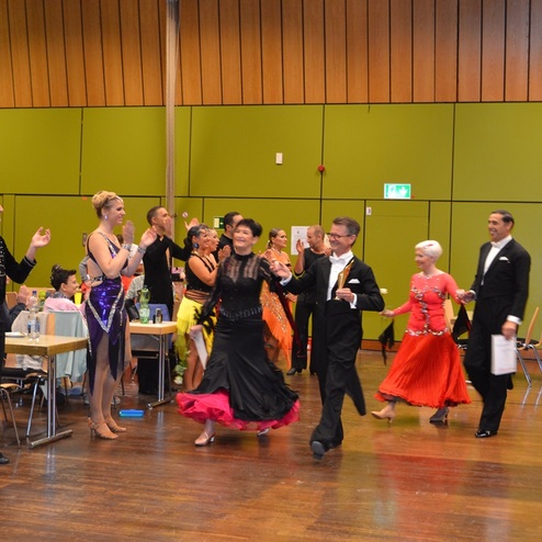 Tanzpaare in festlicher Kleidung betreten die B&uuml;hne, w&auml;hrend Zuschauer applaudieren und feiern.