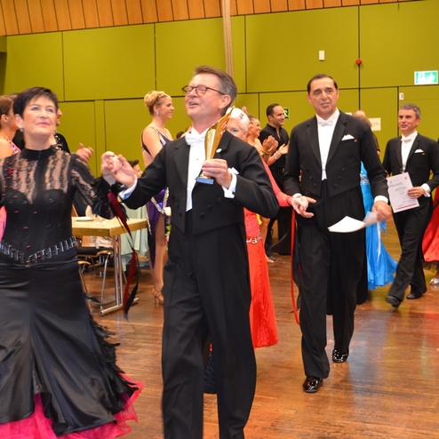 Tanzpaare in festlicher Kleidung beim Wettbewerb, l&auml;cheln und halten Pokale in einem Saal mit Tischen im Hintergrund.